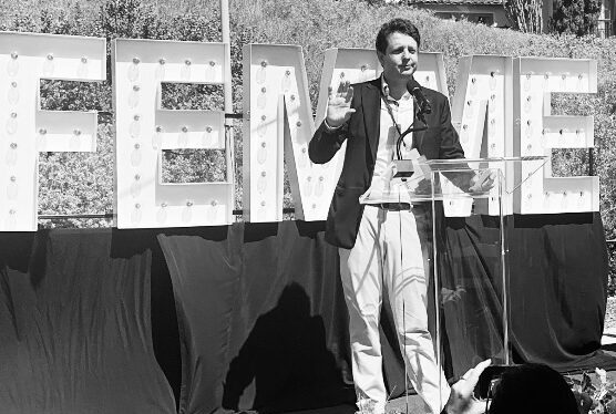 A man speaking at a podium with the large letters "FEMME" behind him